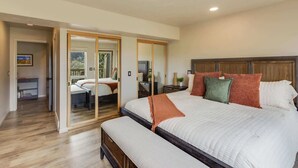 Une chambre avec un grand lit, une tête de lit en bois, un banc, un ventilateur de plafond et une porte coulissante donnant sur une autre pièce.