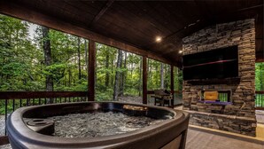 Ein Whirlpool mit Steinwand und daran montiertem Fernseher, umgeben von einer Holzterrasse mit Blick auf einen Wald.