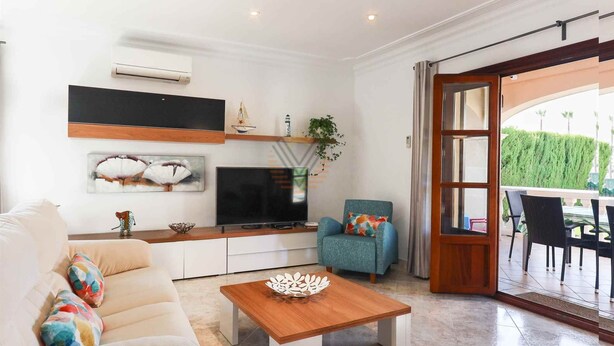 A modern living room with a sofa, coffee table, TV, and a dining area with chairs. - Villa Llac Blau – Family, pool & beach Alcudia (Puerto de Alcudia)