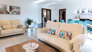 A modern living room with two beige sofas, a wooden coffee table, and a dining area with blue chairs. - Villa Llac Blau – Family, pool & beach Alcudia (Puerto de Alcudia)