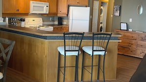 A kitchen with wooden cabinets, a white microwave, a refrigerator, and a bar area with two chairs. - Kimberley Chalet (Kimberley)