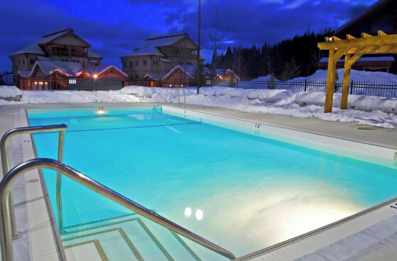 Une piscine à l’eau d’un bleu cristallin, entourée de neige et surmontée d’une pergola en bois.