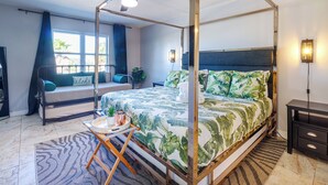 Ein Bett mit Baldachin mit tropischem Blattmuster, ein Deckenventilator, ein Nachttisch mit Lampe und Blick auf Palmen durch das Fenster.