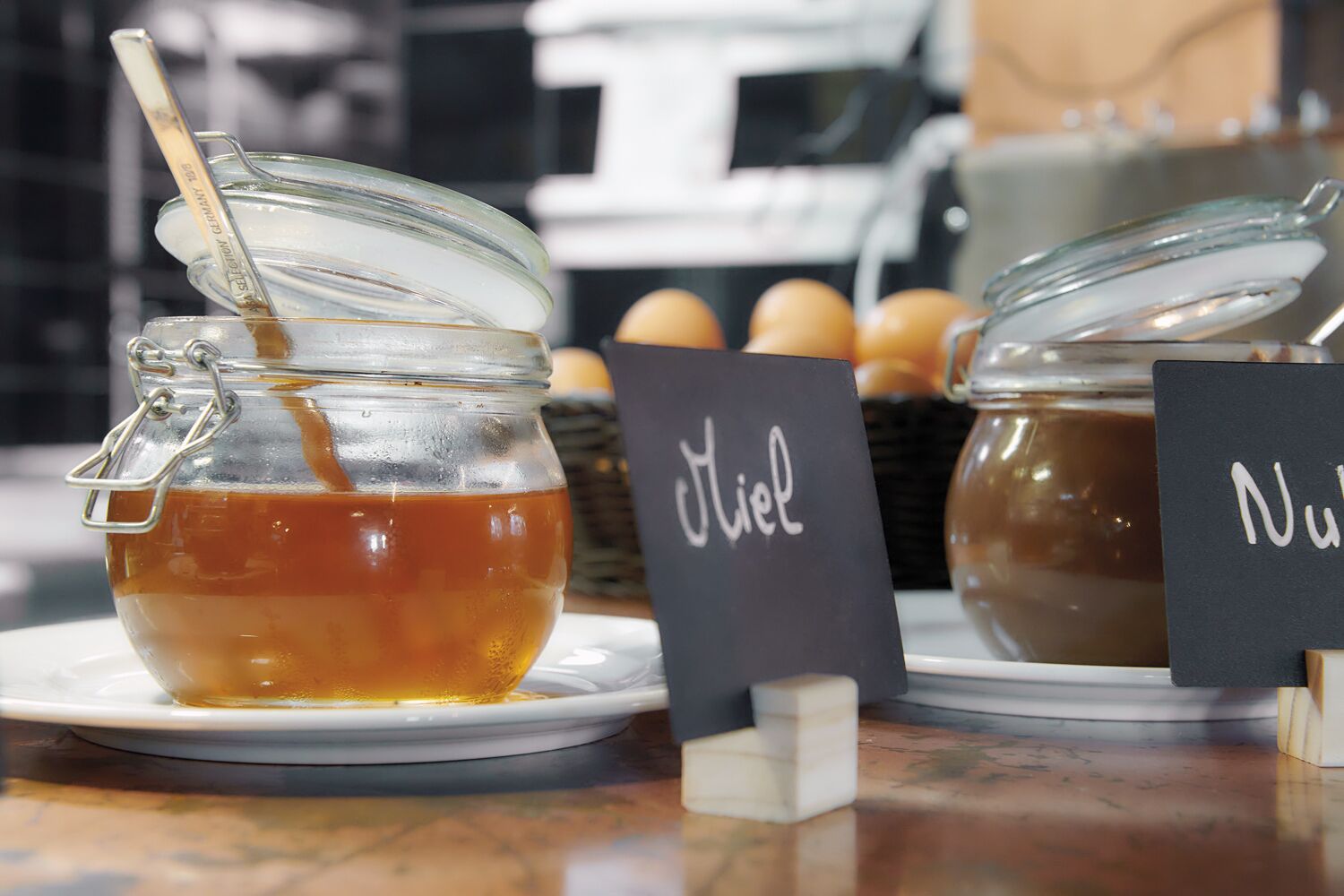 A glass jar with a metal spoon, labeled "Miel," and another jar labeled "Nut."