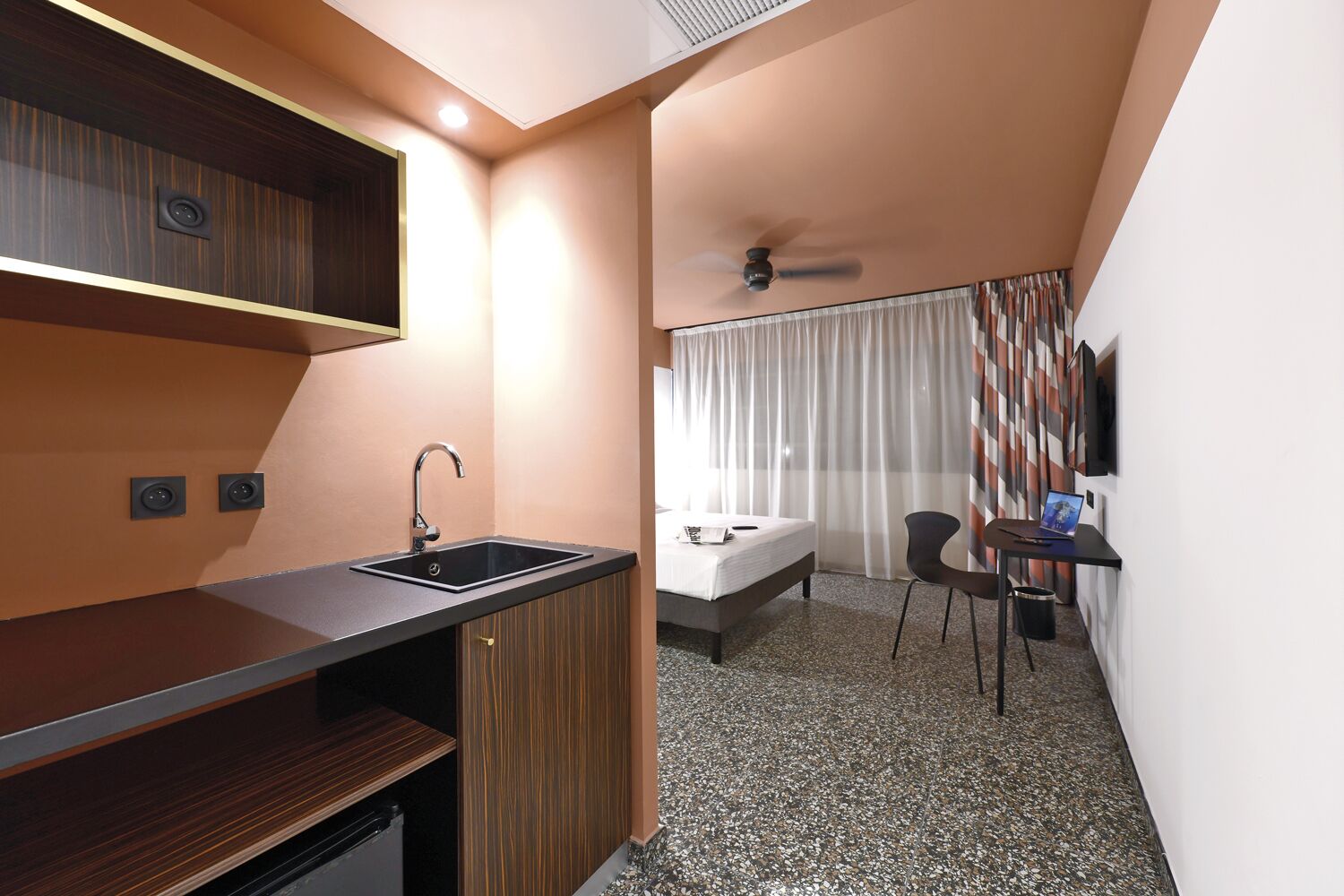 A compact hotel room with a kitchenette, a bed, a desk with a laptop, and a ceiling fan.