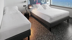 Een hotelkamer met twee bedden, een groot raam met uitzicht op zee, een plafondventilator en een muurschildering.