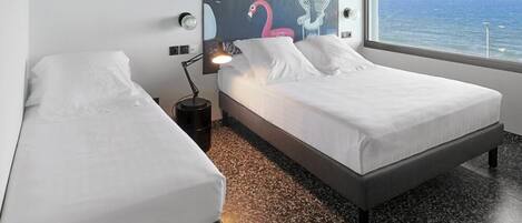 A hotel room with two beds, a large window with a view of the sea, a ceiling fan, and a wall mural.