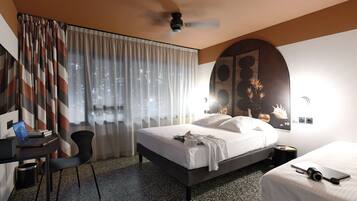 Ein Hotelzimmer mit einem Bett, einem Schreibtisch mit Laptop, einem Sessel, einem weiteren Bett, einem Wand-Fernseher und einem Deckenventilator.