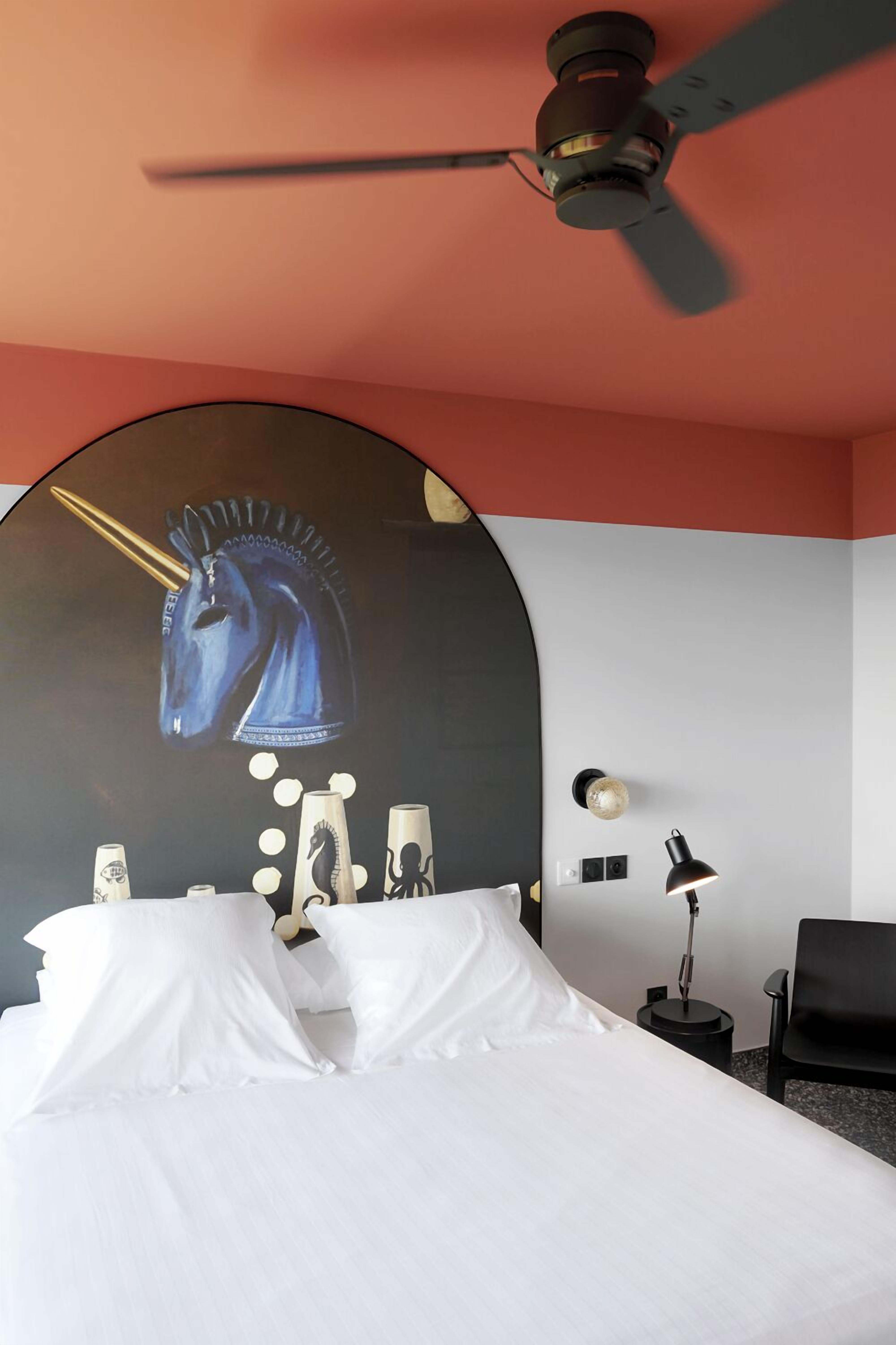 Una habitación de hotel con una cama, un ventilador de techo, una lámpara de pared y una silla negra.