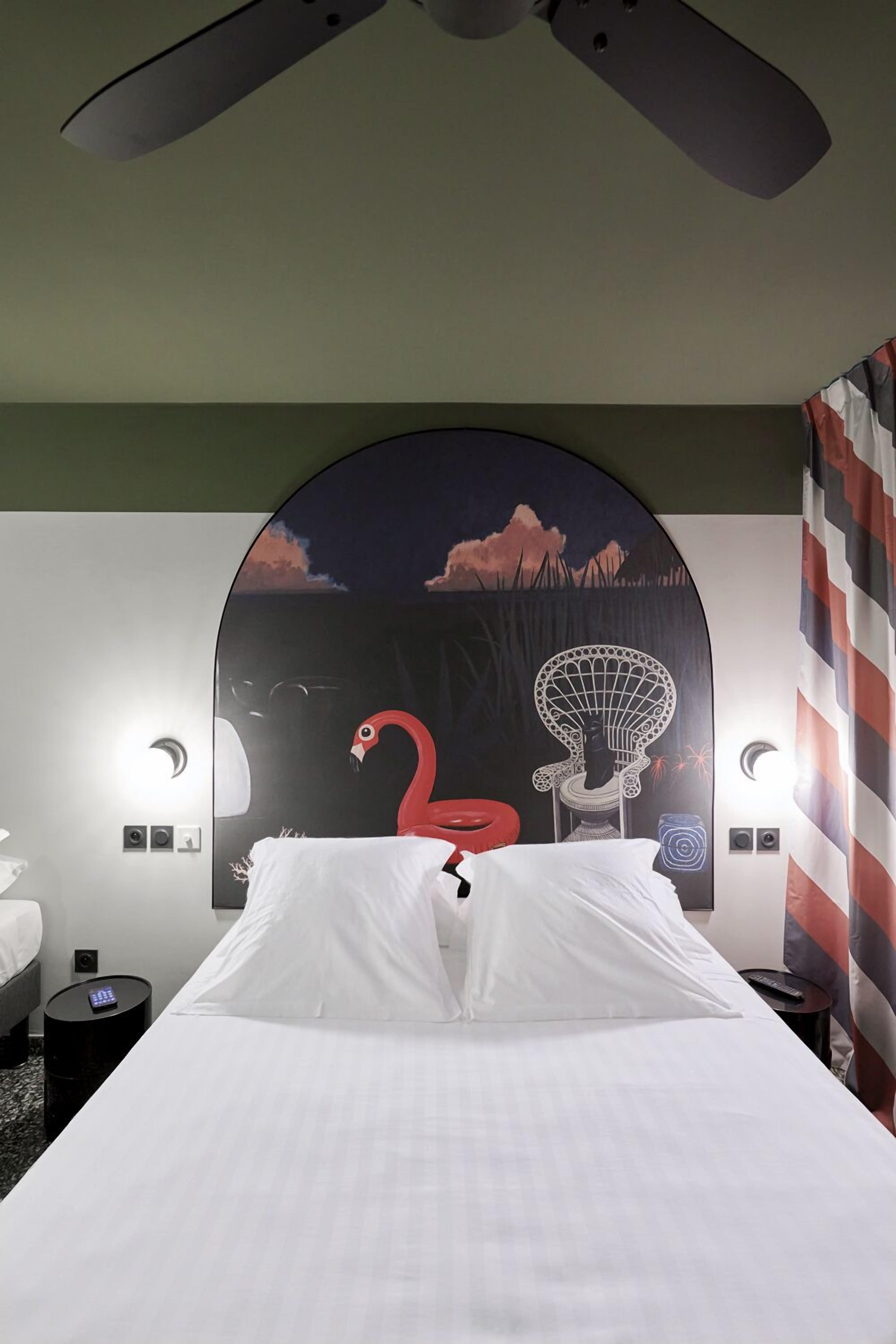A hotel room with a bed, a ceiling fan, a wall mural, a nightstand, and a remote control.