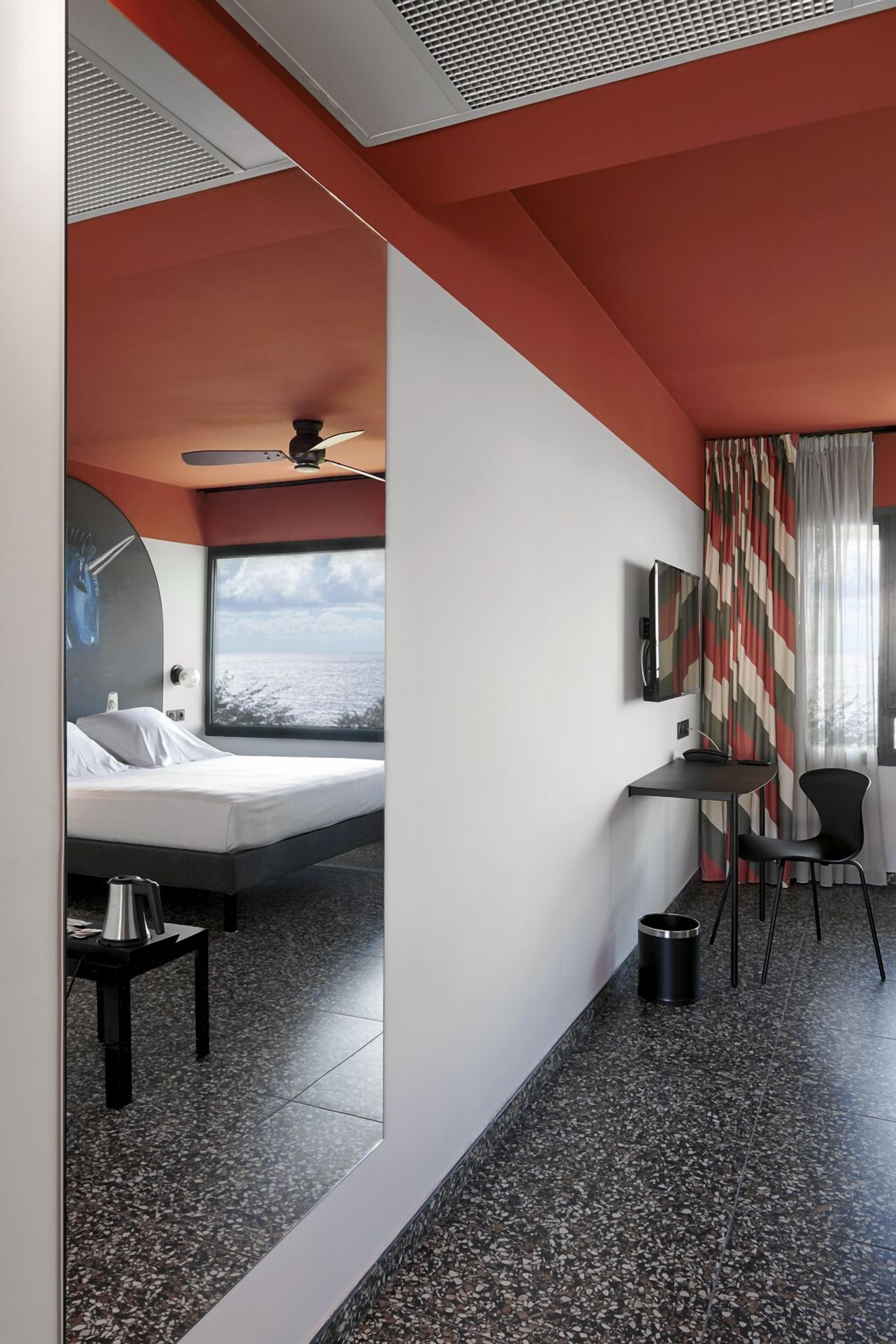 A modern hotel room with a bed, a desk, a chair, a TV, and a large window offering a view of the sea.