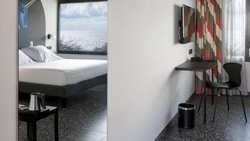 A modern hotel room with a bed, a desk, a chair, a TV, and a large window offering a view of the sea.