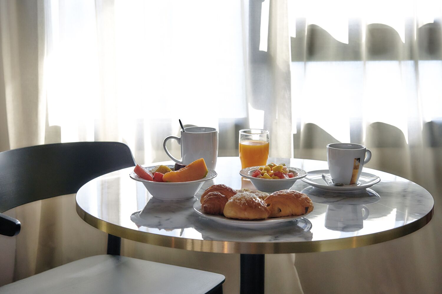 Eixam de matí amb croissants, fruita, un got de suc d'aranja i dues tasses sobre una taula de marbre al costat d'una finestra amb cortines primes.