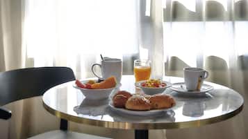 Eixam de matí amb croissants, fruita, un got de suc d'aranja i dues tasses sobre una taula de marbre al costat d'una finestra amb cortines primes.