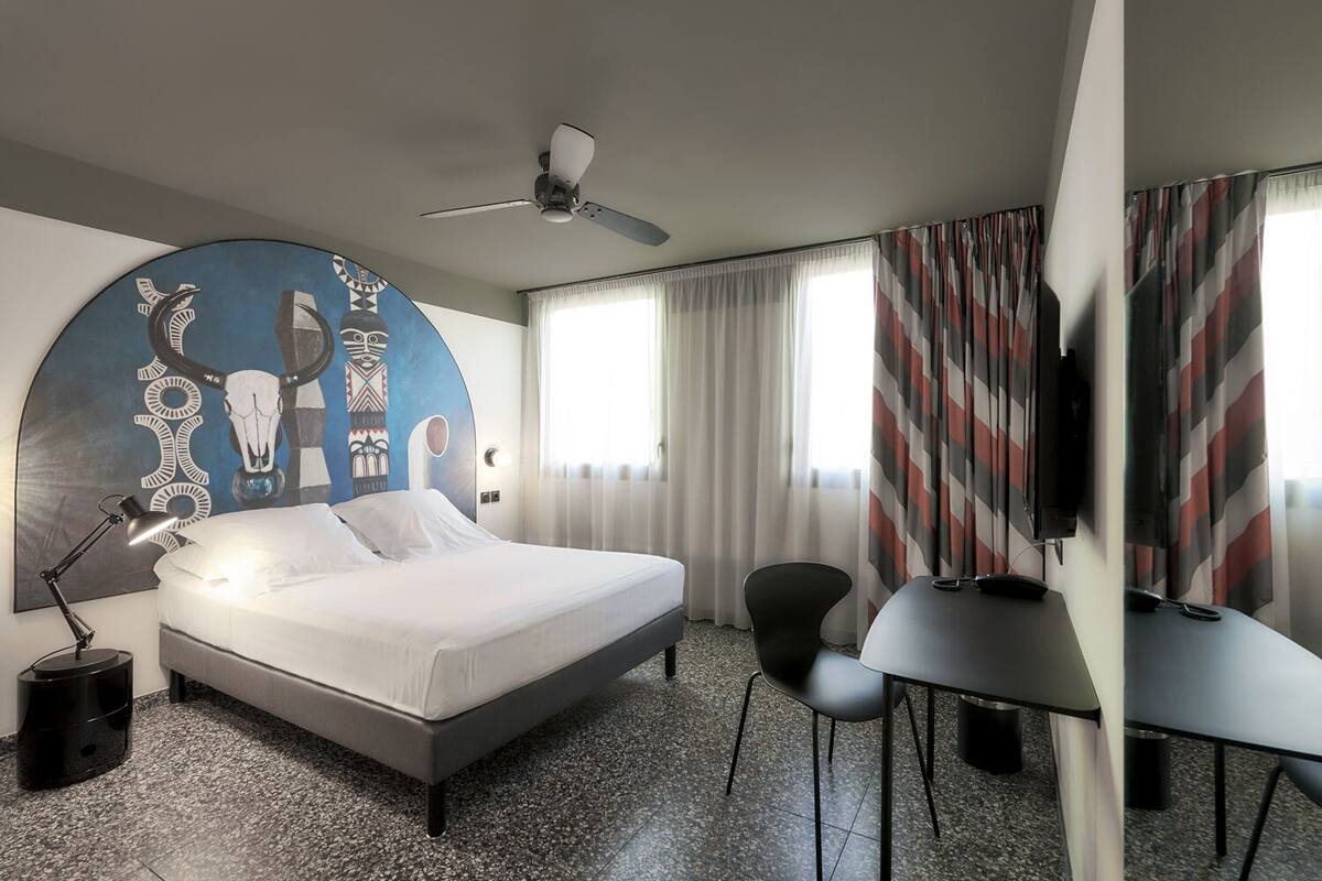 Een hotelkamer met een bed, een bureau, een stoel, een plafondventilator en een groot wandtapijt.