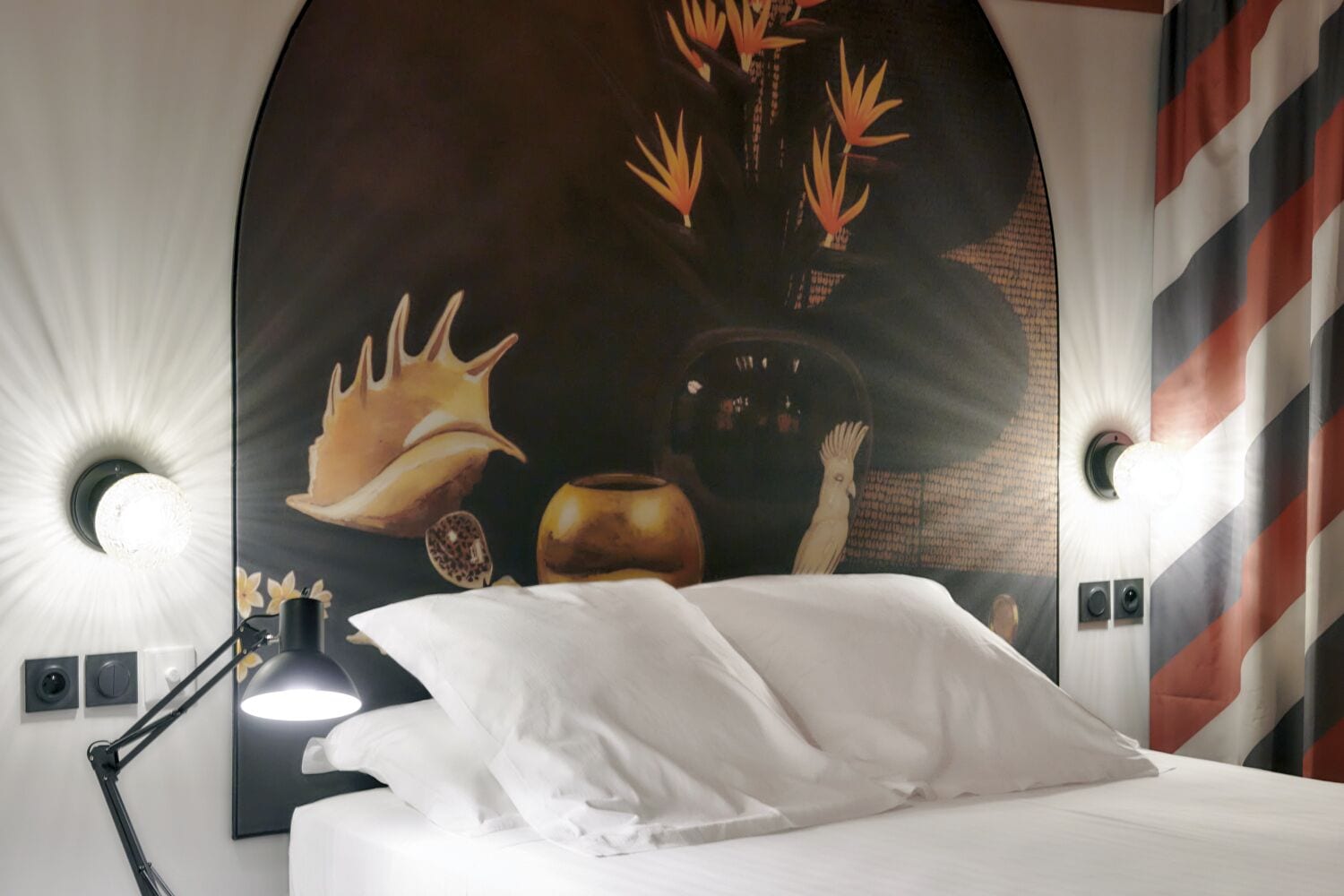 Hotelkamer met een bed, nachtkastje met lamp, kunstwerk aan de muur met een vogel en een schelp, en een gestreept behang.