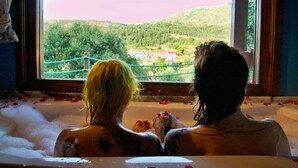 Two people in a bathtub with rose petals, looking out at a scenic view of a village and hills. - Toca dos Lobos - Ecological Cabin (Caminha)