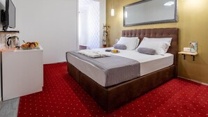 Een hotelkamer met een groot bed, een televisie, een klein tafeltje met een vaas en fruit, en een rode vloerbedekking.