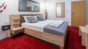 A hotel room with a wooden bed, a bedside table with a lamp, a mirror, and a red carpet. - Deluxe Room in Hotel Rose Split (Split)