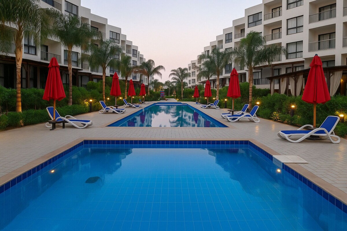 Ein Swimmingpool mit Liegestühlen und roten Sonnenschirmen, umgeben von Palmen und modernen Gebäuden.