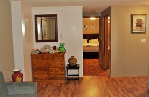 A hallway with a wooden dresser, a vase, and a mirror. There is a bed with a red rug in the background.