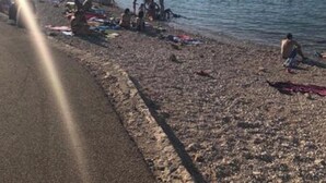 Ranta, jossa ihmiset uivat ja ottavat aurinkoa, kirkas taivas ja taustalla näkyvä rakennus.