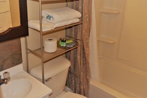 A bathroom with a toilet, a sink, a shelf with towels, toilet paper, and toiletries.