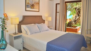 A neatly made bed with a blue blanket, two bedside lamps, a framed painting on the wall, and a view of a tree through an open door. - Holiday House Angelos on Agios Gordios Beach (Corfu)