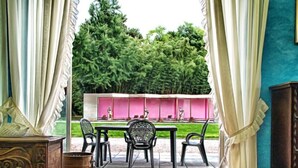 Um quarto com piso de madeira, tapete estampado, uma mesa com cadeiras, um armário e vista para um jardim com uma parede rosa e estátuas.