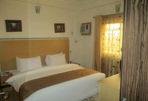 A hotel room with a bed, bedside table, lamp, and a window with curtains. - Room-abuja Elomaz Hotel-guest Suite (Abuja)