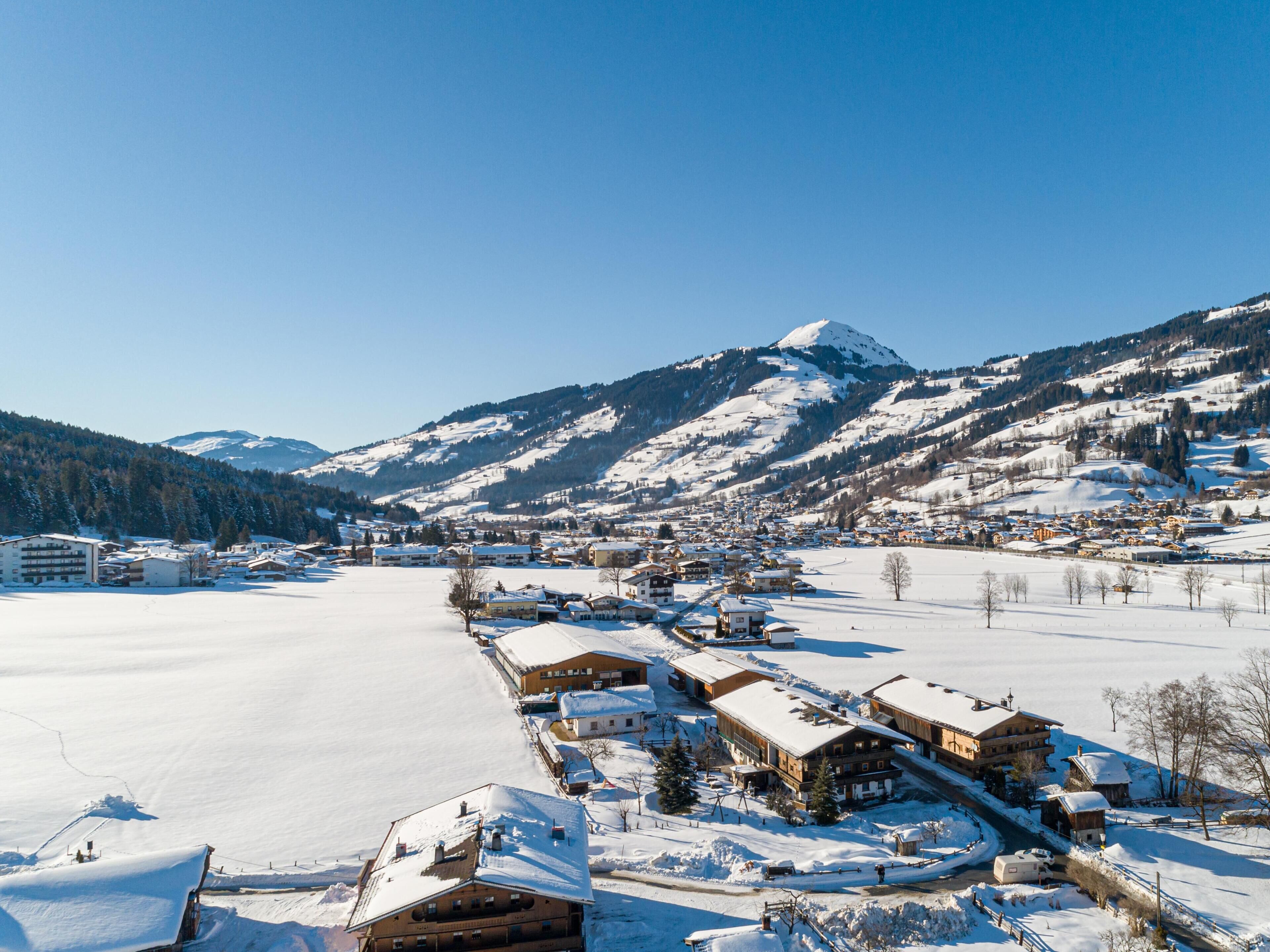 A snow-covered village with buildings along a frozen lake, surrounded by mountains.