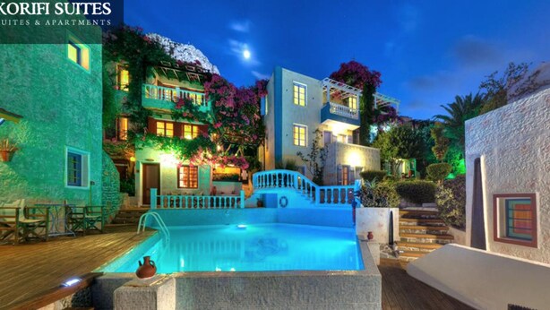 A pool area with a stone deck, a small fountain, and a staircase leading to a building with balconies and a garden. - Very Spacious and Well Equipped Room near the Sea (Piskopiano)