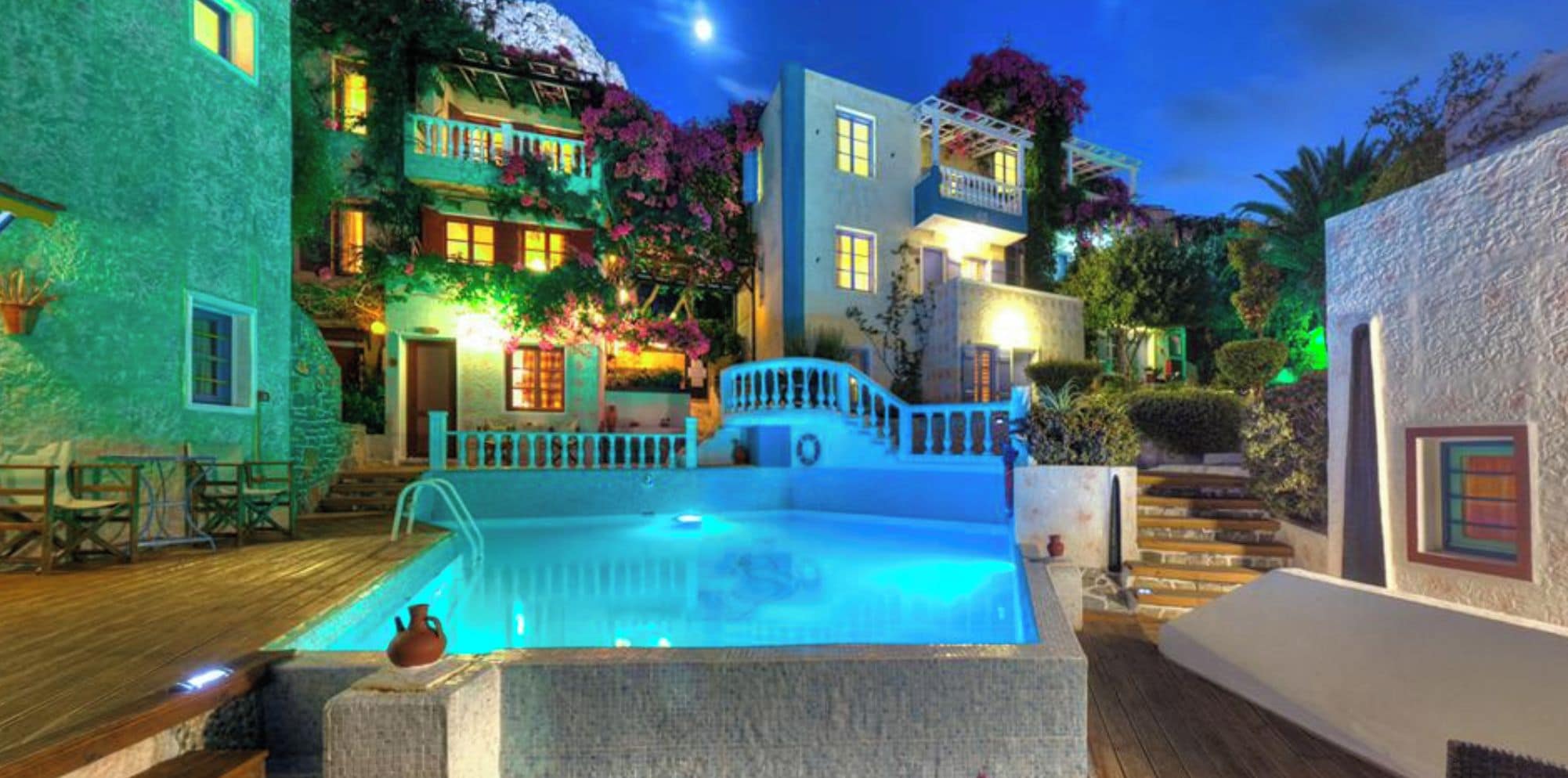 A pool area with a stone deck, a small fountain, and a staircase leading to a building with balconies and a garden.