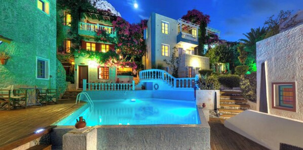 Un área de piscina con un deck de piedra, una pequeña fuente y una escalera que lleva a un edificio con balcones y un jardín.