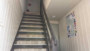 Eine Treppe mit Holzstufen und Metallgeländern, die zu einer Zwischenebene führt, auf der sich ein Feuerlöscher und eine Wand mit verschiedenen Plakaten befinden.
