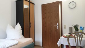 A bedroom with a bed, a wardrobe, a door, a wall clock, a table with a chair, and a potted plant. - Einzelzimmer - Pension Pizzeria Verona (Großräschen)