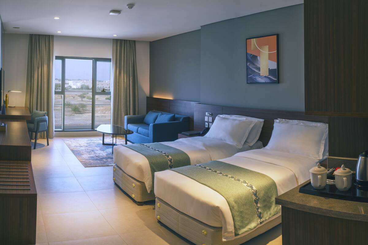 A hotel room with two beds, a desk, a chair, a sofa, and a large window with curtains.