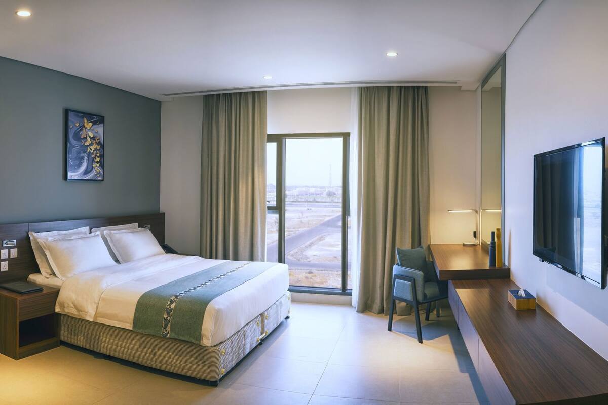 A modern hotel room with a bed, desk, chair, and large window.