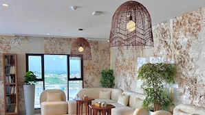 A spacious living room with a large sofa, a coffee table, and a bookshelf. There are two hanging lights and a large window providing a view of the city. - Boulevard hotel (Seeb)