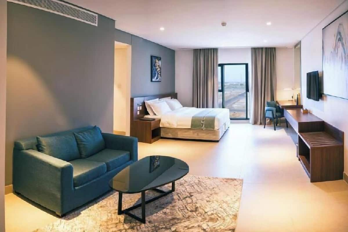 A modern hotel room with a bed, a sofa, a coffee table, a desk, and a large window.