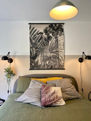 Un dormitorio con cama, un cuadro de hojas tropicales en la pared, dos lámparas de noche y una lámpara colgante.