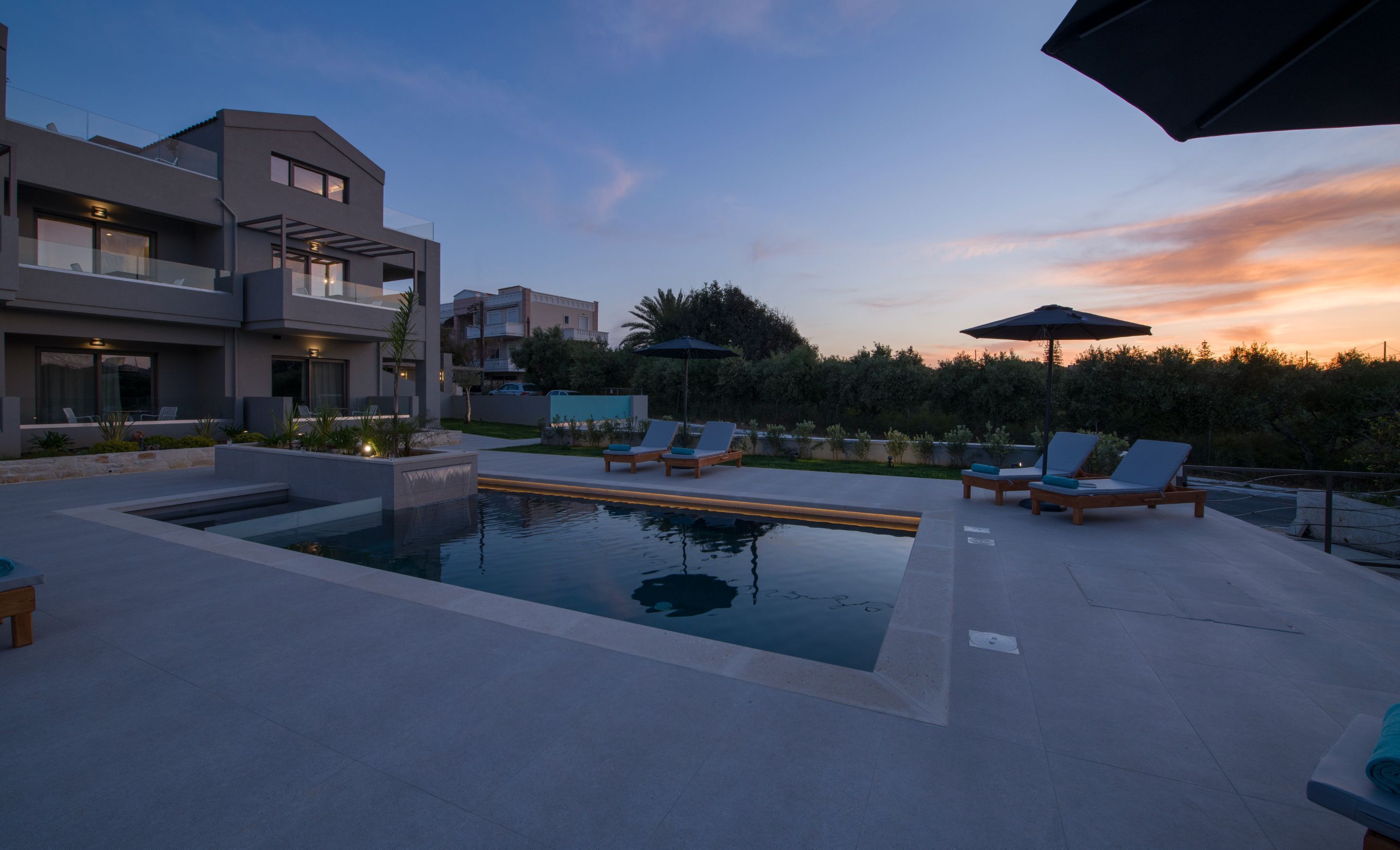 Un área de piscina exterior moderna con sillones de descanso y sombrillas, rodeada de edificios contemporáneos.