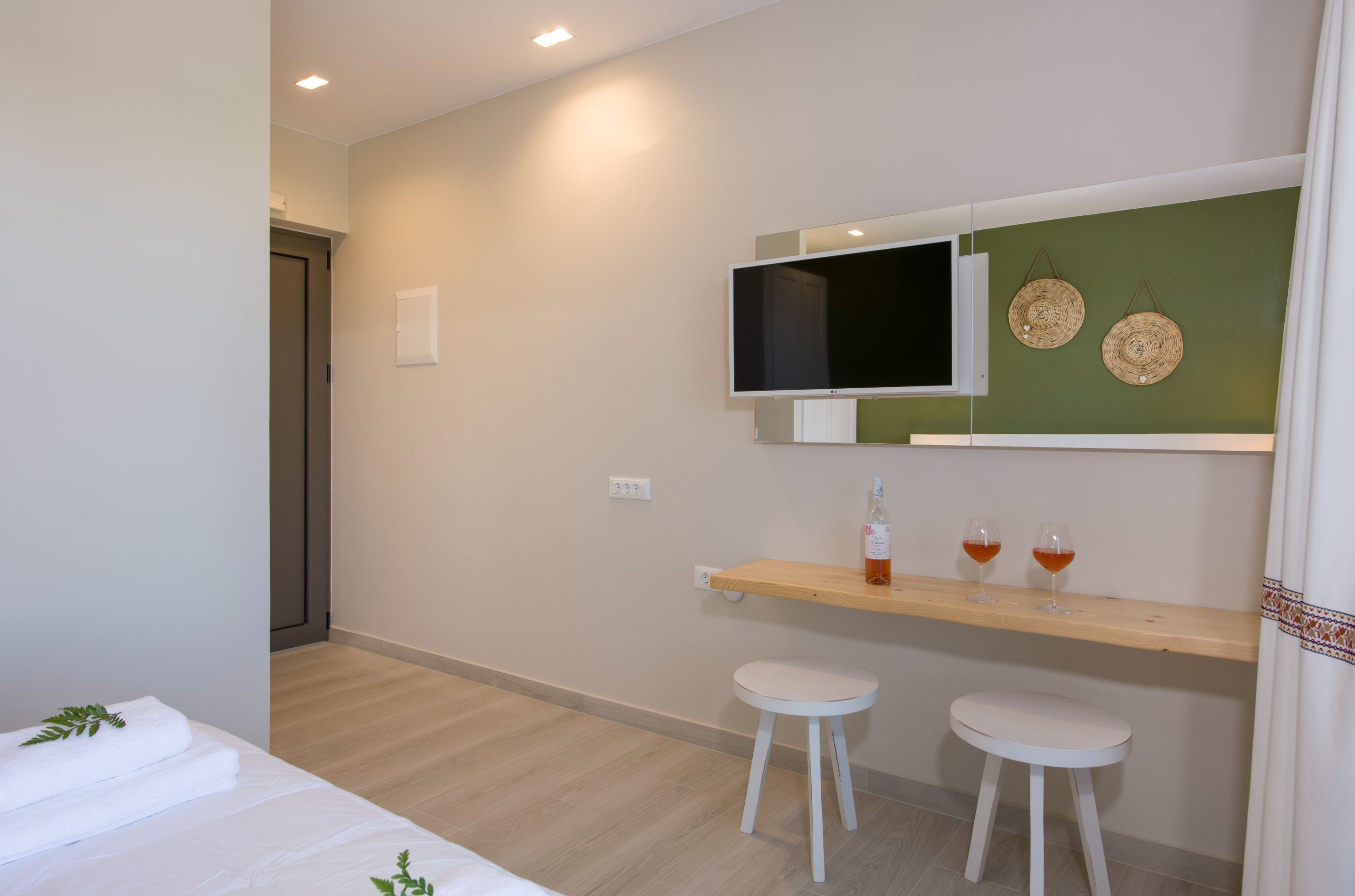 Ein modernes Hotelzimmer mit einem Bett, einem an der Wand befestigten Fernseher, einer kleinen Holzablage mit Gläsern und einer Flasche sowie zwei weißen Hocker.