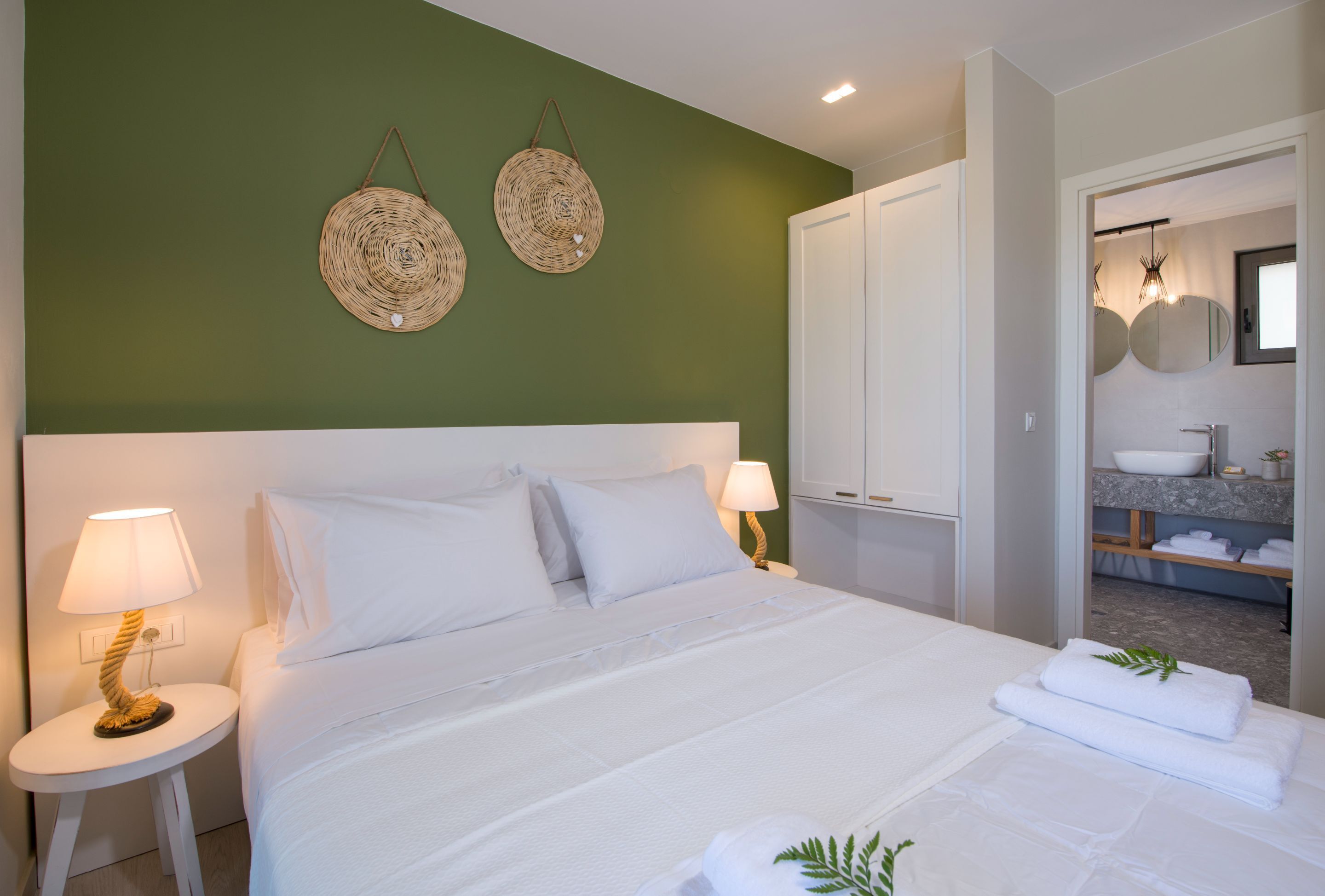 A hotel room with a large bed, two bedside lamps, a green accent wall, and a bathroom visible through an open door.