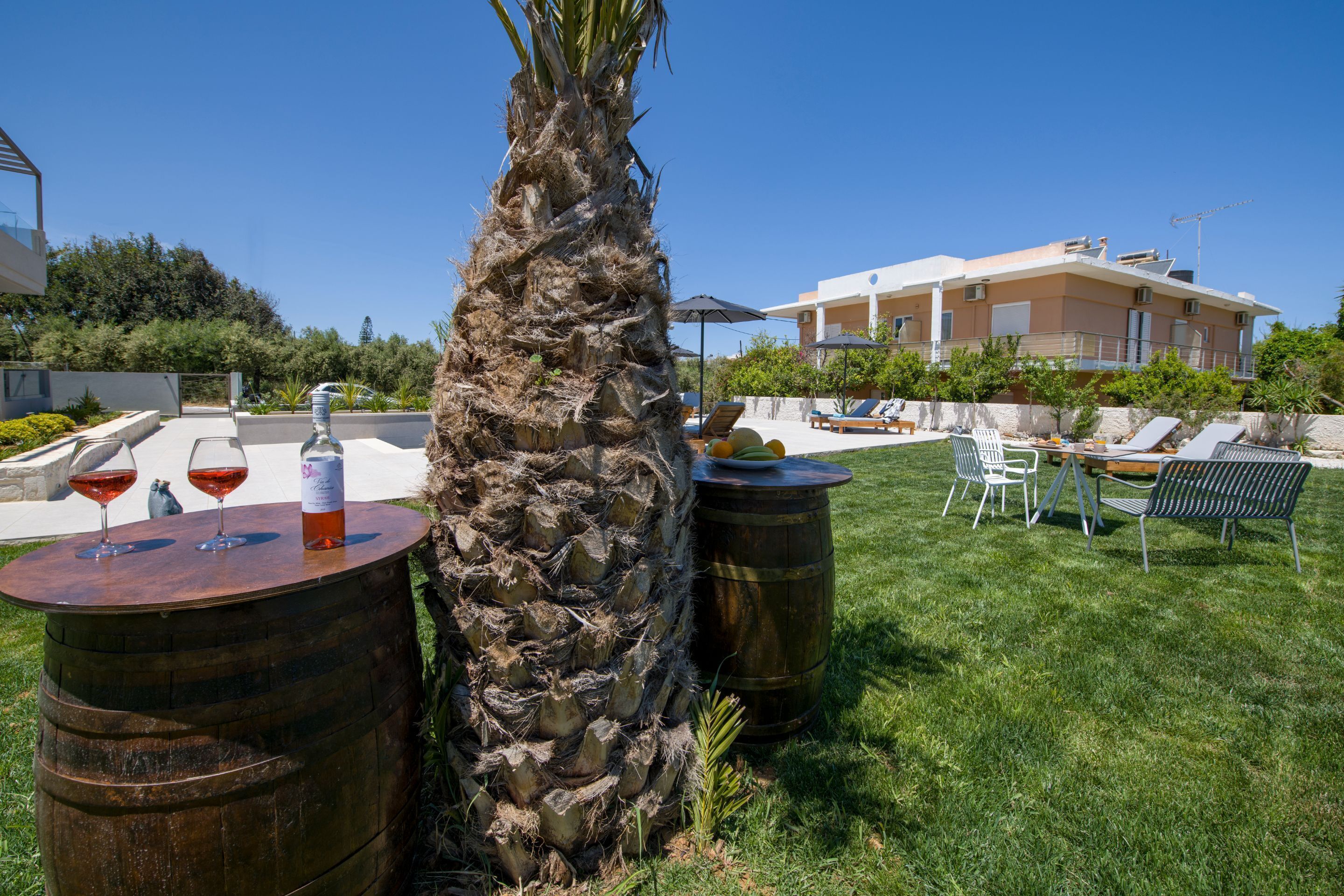 A tropical outdoor area with a palm tree, wooden barrels, and a poolside lounge with chairs and umbrellas.