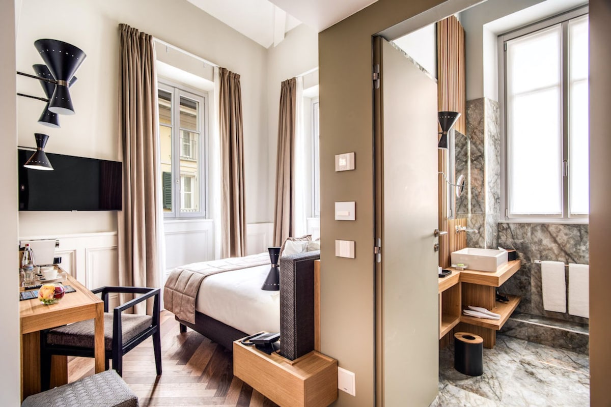 A hotel room with a bed, bedside table, chair, desk, and a bathroom with a sink and mirror.