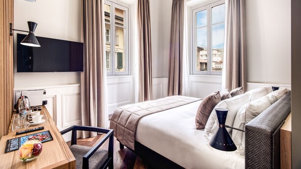Una habitación de hotel con cama, mesita de noche, lámpara, cortinas y televisor montado en la pared.
