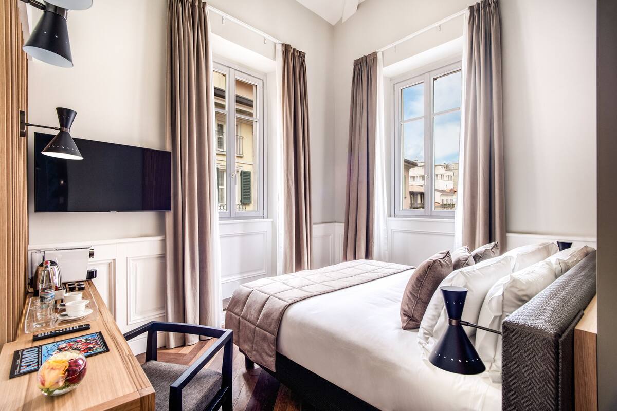 Een hotelkamer met een bed, nachtkastje, lamp, gordijnen en een aan de muur bevestigde televisie.