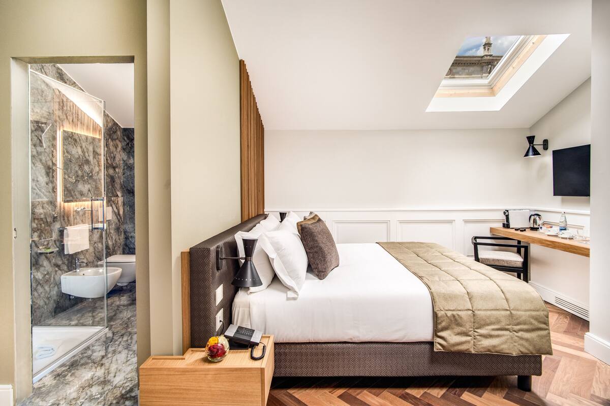 Een moderne hotelkamer met een groot bed, een nachtkastje met een telefoon en een schaal fruit, een bureau met een stoel, een badkamer met een marmeren wand en een wastafel, en een dakraam boven het bed.