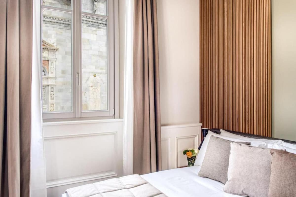 Habitación de hotel con cama, ventana con cortinas, pared de paneles de madera y vista a un edificio.